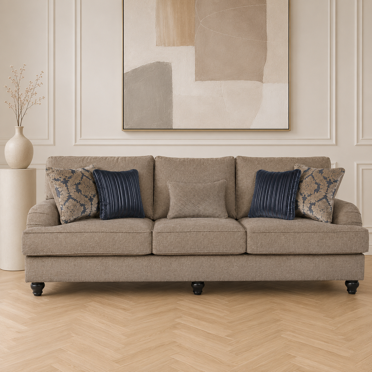 A four
-seat upholstered sofa in a warm taupe-beige fabric, styled with two navy blue ribbed pillows and two beige-and-navy patterned accent pillows. The sofa is set in a bright living room with a light wood herringbone floor, a white side table with a vase, and a large abstract wall art piece above.