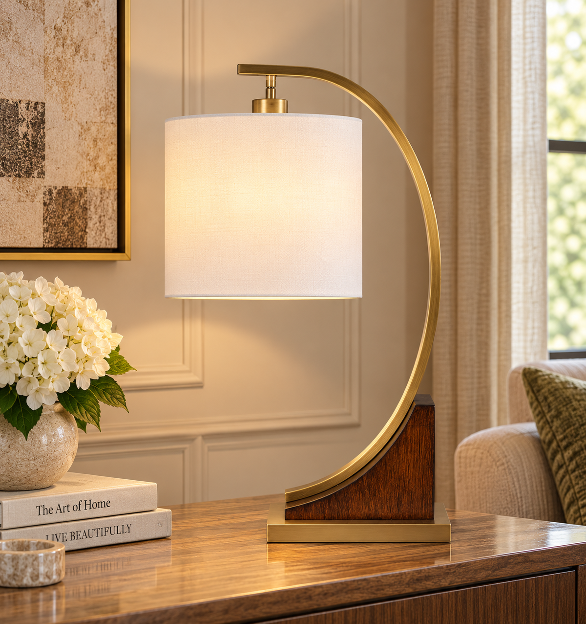 A modern table lamp with a curved gold metal frame, a white fabric drum shade, and a dark wood base, shown lit on a wooden console table in a warm, elegant room.