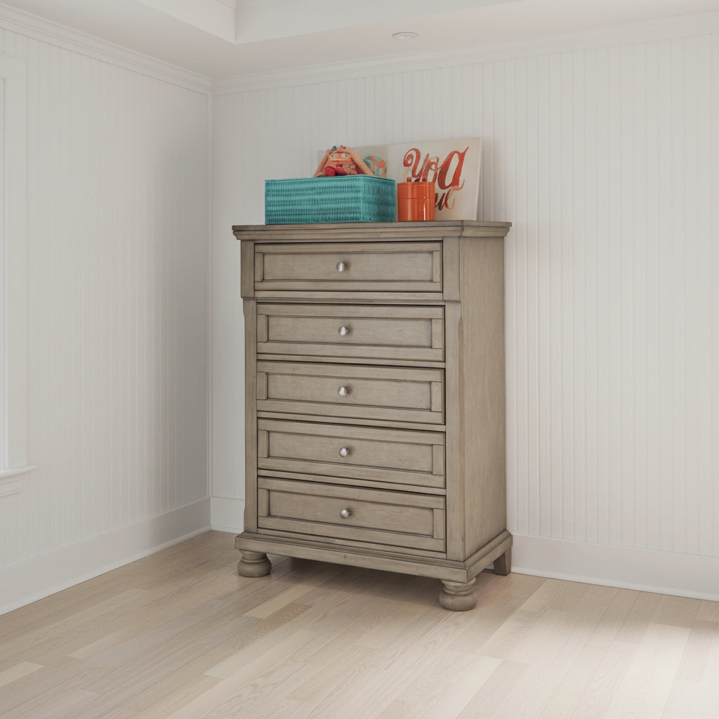A tall, five-drawer chest of drawers in a distressed light gray-brown wood finish, featuring small round silver knobs and turned feet, shown in a room setting with a basket and orange cups on top.