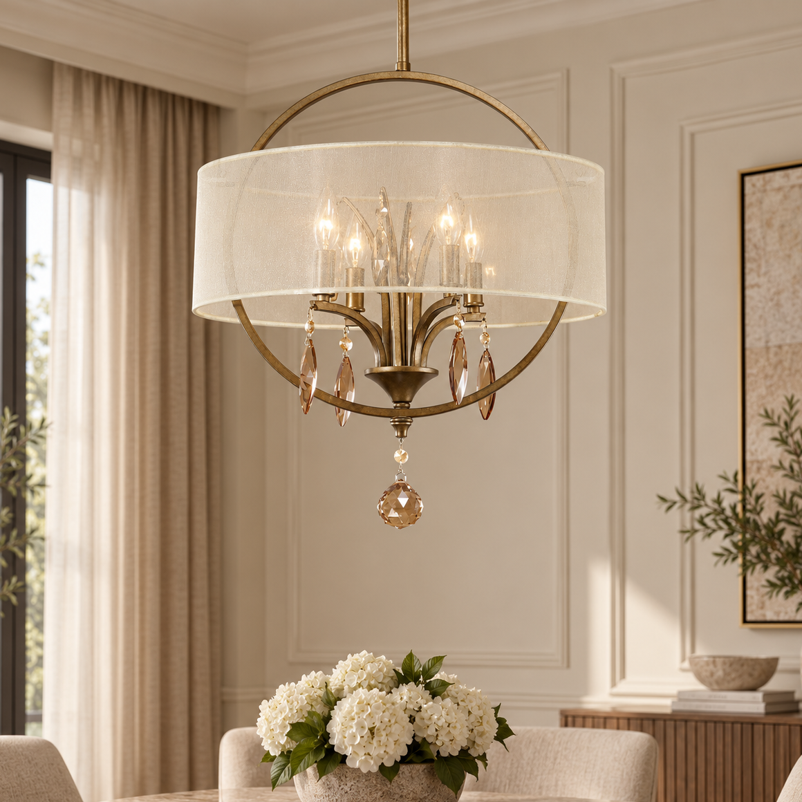 A brass and white drum chandelier with hanging crystal accents is suspended over a dining table in a beautifully decorated room with beige walls, curtains, and a floral centerpiece.