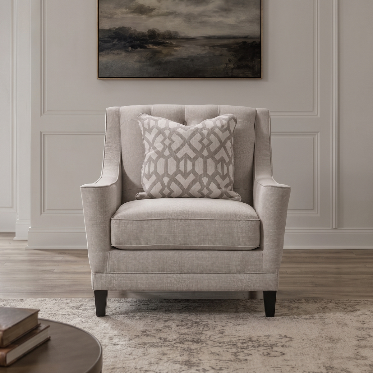 A light beige upholstered armchair with a gray and white geometric patterned throw pillow, set in a bright, modern living room with a framed landscape painting on the wall.