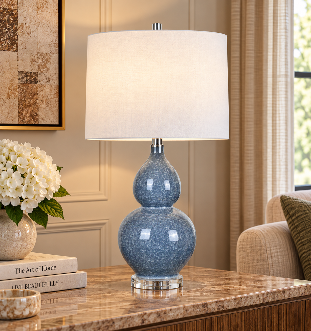 A table lamp with a glossy blue ceramic double-gourd base and a white fabric drum shade, placed on a marble side table in a styled living room setting.